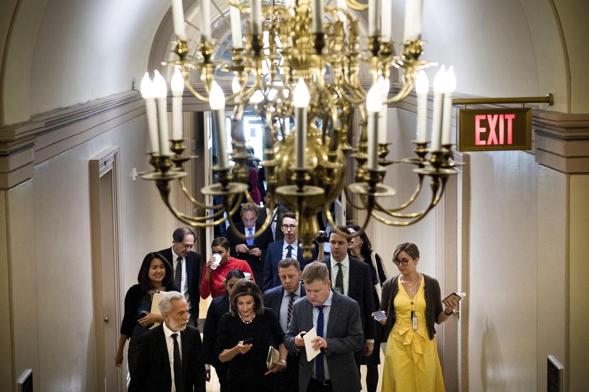 Speaker of the House Nancy Pelosi walks from a meeting with House Democrats. The photograph has a huge chandelier in the foreground.