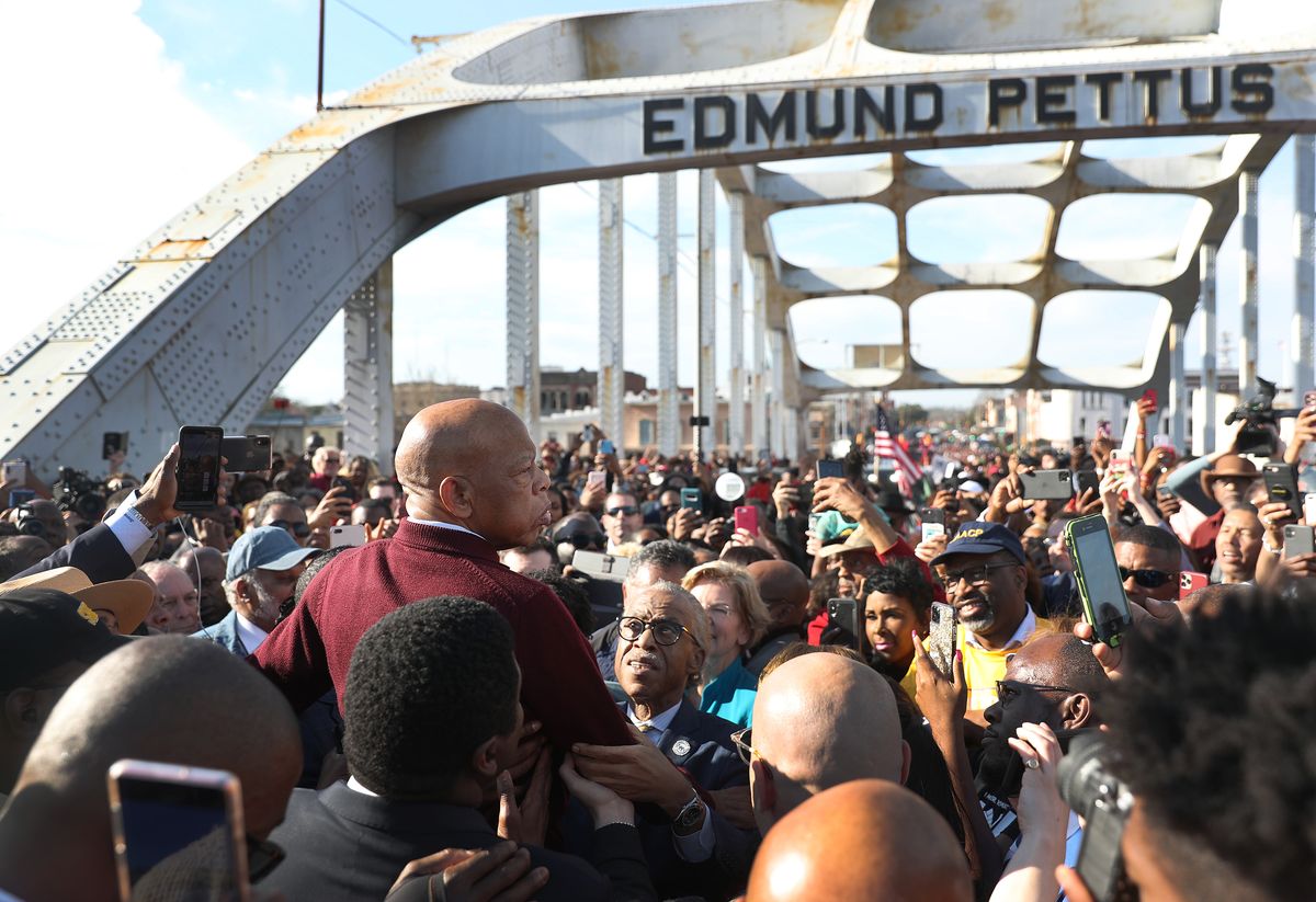 Lewis, is a red sweater, is raised above a crowd packing the Edmund Pettus Bridge. Many raise cellphones to capture the moment. Lewis looks to his right, his face grave.