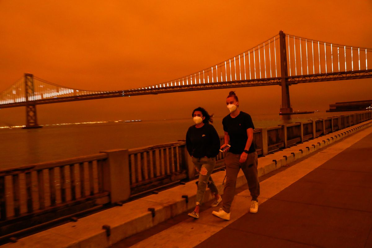 People pass by the Bay Bridge as dark orange skies hang over downtown San Francisco, Calif. Wednesday, September 9, 2020 due to multiple wildfires burning across California and Oregon.